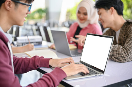 Close-up image of a smart young Asian male college student browsing internet, using laptop, scrolling on a website while working on a group project with his friends. laptop white screen mockupの写真素材