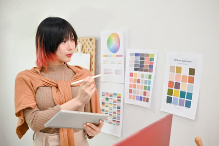 Attractive and focused young Asian female graphic designer working on her new design project in her studio, using tablet and choosing color from color swatch sample.の写真素材