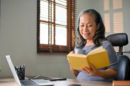 Happy and relaxed Asian-aged businesswoman is reading a book and sipping her morning coffee at her desk.の写真素材