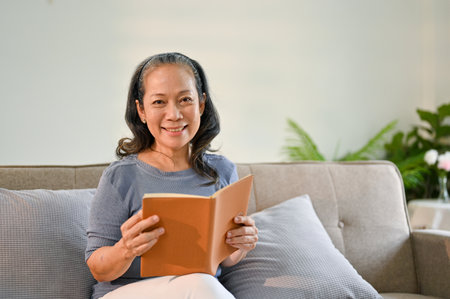 Attractive and happy Asian-aged woman sits on a sofa in her minimal and cozy living room with a book in her hands.の写真素材