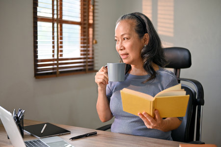 Happy and relaxed Asian-aged businesswoman is reading a book, sipping her morning coffee and daydreaming about happiness at her desk.の写真素材