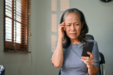 Confused and unhappy Asian-aged woman sits at her desk having a problem with her internet connection on her smartphone, having a headache with technology.の写真素材