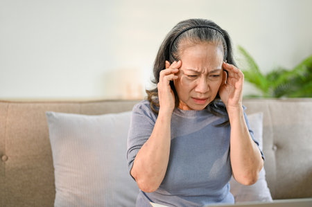 Stressed and unwell senior Asian woman sits on sofa in her living room, touching her temples and suffering from headache or migraine. Health problems conceptの写真素材
