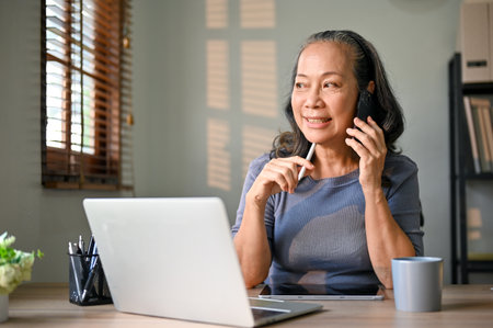 Successful and happy Asian-aged businesswoman is talking on the phone with her business partner while working in her office.の写真素材