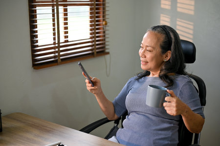 Happy and relaxed Asian-aged businesswoman sipping coffee and using her phone at her desk.