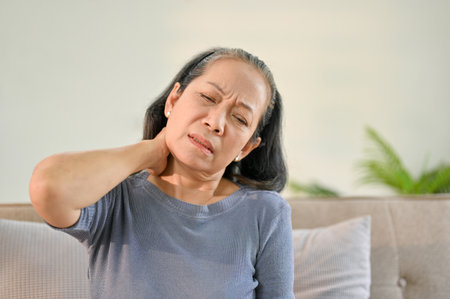 Unhappy senior Asian woman sits on sofa in her living room suffering from neck pain and rubbing her neck.の写真素材