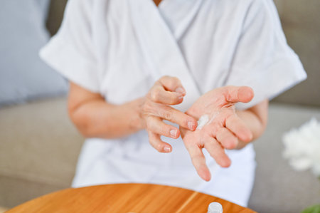 Close-up image of an Asian-aged woman in bathrobe rubbing her face serum on her hand, applying...