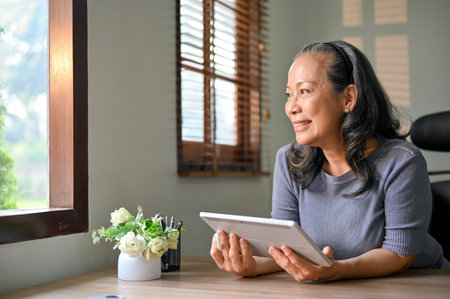 Charming and smiling Asian-aged businesswoman sits at her desk with her tablet and looks out the window, daydreaming about her retirement happiness.の写真素材