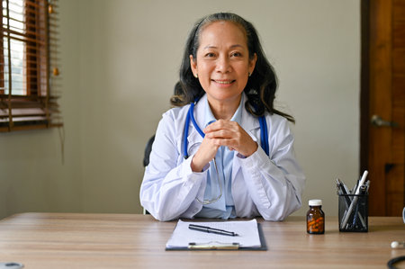 Portrait of a professional Asian-aged female doctor in white gown sits at her desk in the hospital office. health care business and people conceptの写真素材