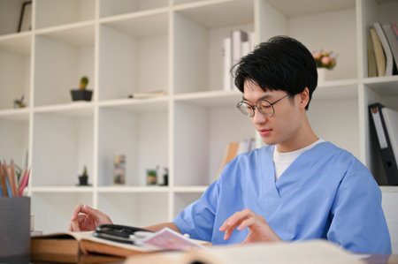 Smart young Asian male medical student in a uniform focuses on reading a book, preparing for exam, sitting in the medical school library. medical students conceptの写真素材