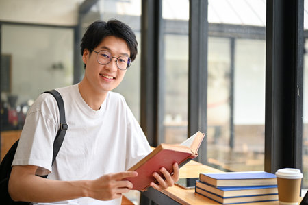 Smart and handsome young Asian male college student wearing eyeglasses sits in a coffee shop with schoolbooks.の写真素材