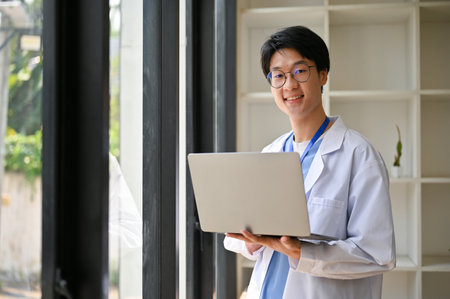 Smart and handsome young Asian male medical student in white gown stands in a hospital corridor with his portable laptop.の写真素材