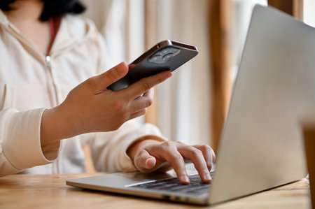 An Asian female college student or freelancer in casual clothes using her smartphone and laptop in a...