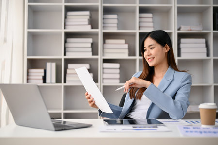 Professional and confident millennial Asian businesswoman reading and reviewing business reports at her desk in her private office.の写真素材