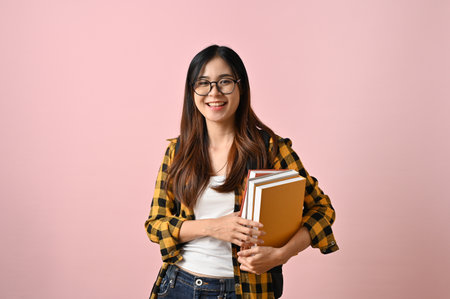 Wise and pretty young Asian female college student in a glasses and flannel shirt holding schoolbooks while standing against a pink isolated background.の写真素材