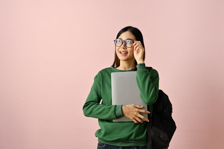 Wise and inspired young Asian female college student touching her glasses, holding her laptop and...