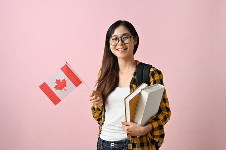 Smiling and pretty young Asian female college student in a flannel shirt with her school books holding a flag of Canada against a pink isolated background. Exchange students conceptの写真素材