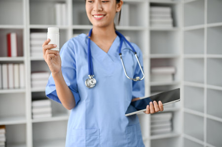 Young female professional doctor holding a bottle of pills while standing in her office room and holding tablet in her handの写真素材