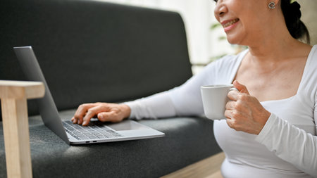 Close-up image of a beautiful and happy senior woman sipping coffee and using her laptop on sofa in...