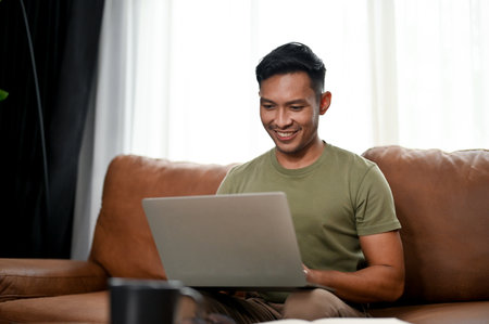 Smart and handsome millennial Asian man using laptop on a sofa in his living room. work from home, free time, leisureの写真素材