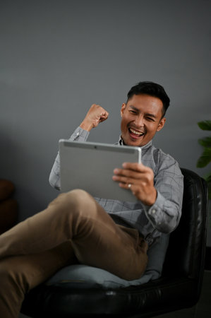Portrait of a cheerful millennial Asian man in casual clothes looking at his tablet screen and celebrating his success.の写真素材