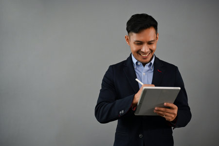 Professional and successful millennial Asian businessman in formal business suit using his digital tablet while standing on an isolated gray background.の写真素材
