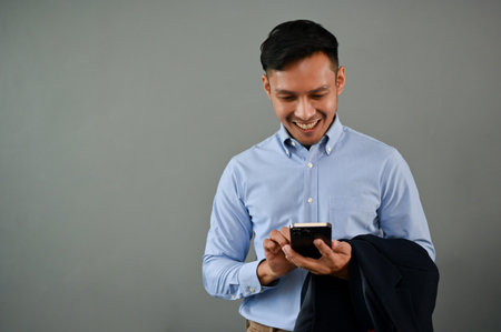 Happy millennial Asian businessman using his smartphone while standing on an isolated gray background. chat, text, message, sms, email, social mediaの写真素材