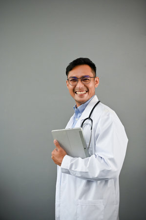 Portrait of a smart and friendly millennial Asian male doctor in a uniform with a stethoscope is holding a digital tablet while standing on an isolated grey background.の写真素材