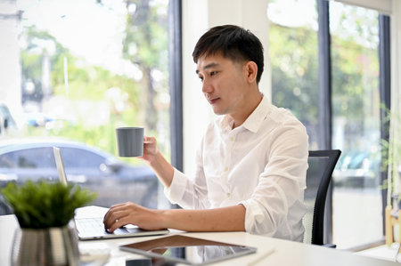 Handsome and smart millennial Asian businessman or male boss in white shirt using his laptop...