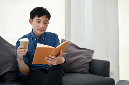 A happy and relaxed millennial Asian man sipping coffee and reading book on a sofa in the living room. Leisure conceptの写真素材