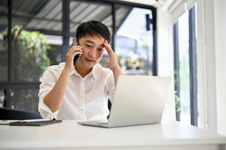 Stressed and thoughtful millennial Asian businessman dealing business with his business client over the phone while sitting at his desk.の写真素材