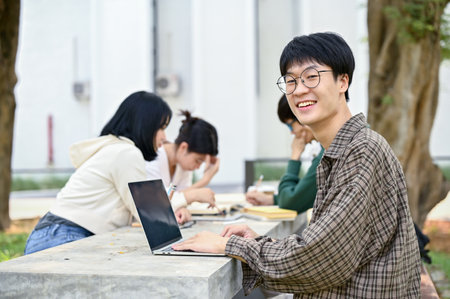 Smiling young Asian male college student in casual clothes with his laptop computer sits at a table in a campus park with his friends.の写真素材