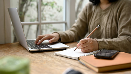 Cropped image of a young Asian woman in cozy sweater taking notes on her notebook and using her...