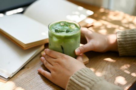 Close-up image of a female in comfy sweater holding a glass of iced matcha green tea at a table in a beautiful minimal cafe.の写真素材