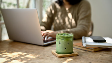 Close-up image of a glass of iced matcha green tea on a wooden table in a minimal cafe with a female sitting behind.の写真素材