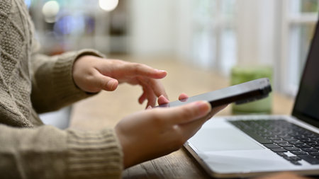 Close-up side view image of a woman in cozy sweater using her smartphone while working on her tasks...