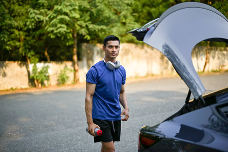 Handsome and sporty millennial Asian man in sportswear stands behind his car, open his car trunk to...
