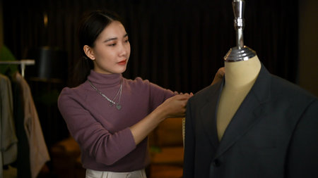 Professional Asian female fashion designer or tailor measuring her suit model on a mannequin with measuring tape, working in her studio.の写真素材