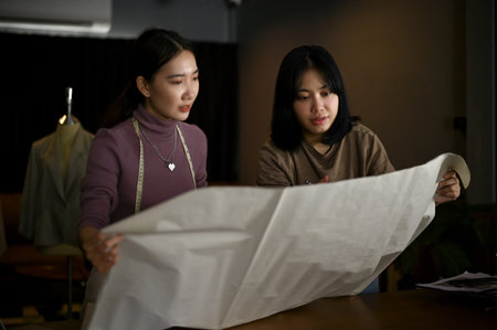 Two Asian female dressmakers or fashion designers are discussing, checking the quality of fabric for their new collection and working in the studio together.の写真素材