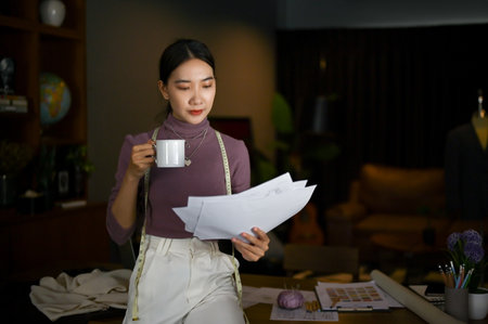 Focused millennial Asian female tailor or fashion designer sipping coffee and checking her model...