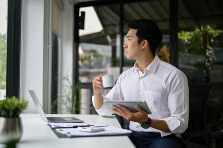Successful millennial Asian businessman or male boss pondering, thinking something while looking out the window and sipping coffee at his desk.の写真素材