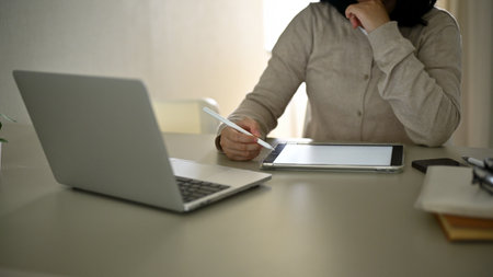 An Asian woman in casual clothes using her digital tablet, working in her home office, sitting at...