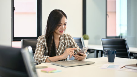 Beautiful young Asian female office worker using her smartphone while sitting in an office co-working space.の写真素材