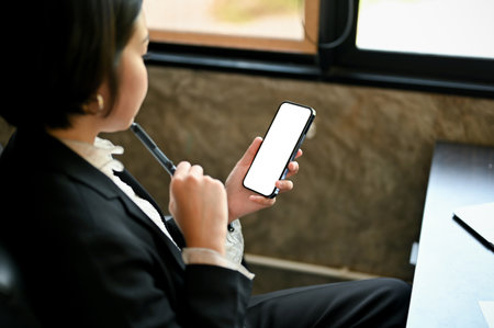Thoughtful millennial Asian businesswoman looking at her phone screen, pensively thinking something while reading a message on her phone at her desk. Smartphone white screen mockupの写真素材