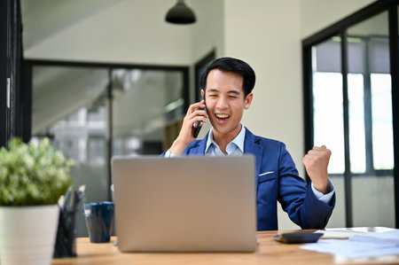 Cheerful and excited millennial Asian businessman showing a clenched fist, getting good news over the phone, and celebrating his success in the office.の写真素材
