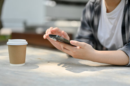 Cropped image of a female in casual clothes using her smartphone while relaxing at a table in a...