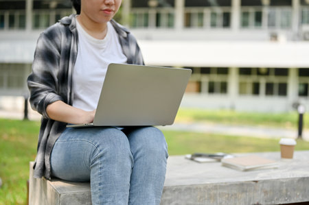 Cropped image of a young Asian female college student in a casual clothes using her portable laptop on a bench in a campus park.の写真素材