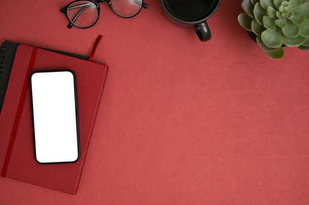 Workspace composition top view with smartphone white screen mockup, red notebook, eyeglasses, coffee cup, potted plant and copy space on red background.の写真素材