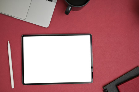 Modern creative office desk top view with digital tablet blank screen mockup, stylus pen, laptop, and coffee cup on red tabletop background.の写真素材