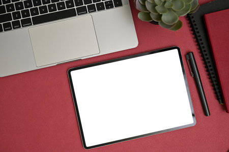 Modern office desk top view with digital tablet white screen mockup, pen, laptop computer, books and succulent on red tabletop background.の写真素材